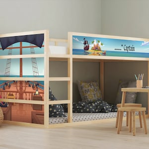 Può includere: Un letto a castello in legno con illustrazioni a tema pirata. Il letto presenta una scena di nave e una scena di isola con la parola "Captain". Il letto è circondato da una cornice in legno chiaro, con un tavolino e sgabelli nelle vicinanze.