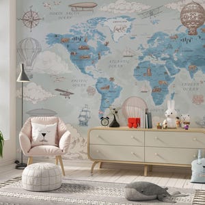 May include: A wall mural featuring a world map in blue and grey tones with illustrations of hot air balloons, ships, and animals. The room includes a light pink armchair with a white bear pillow, a light wood dresser, and a grey whale-shaped cushion.