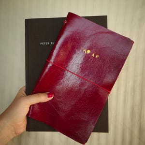 Peut inclure: Un journal en cuir bordeaux avec des lettres dorées et une fermeture élastique rouge. Le journal est posé sur un livre marron foncé avec le texte "PETER ZU". Le journal porte les initiales "AF" en or.