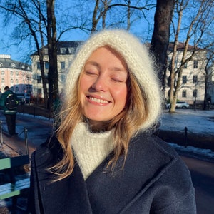 May include: A woman wearing a white knitted hooded scarf and a dark blue coat. She is smiling and has her eyes closed.