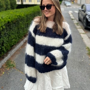 Könnte beinhalten: Eine Frau trägt einen flauschigen, weiß-marineblauen gestreiften Pullover mit langen Ärmeln und einen weißen Rock. Sie trägt eine Sonnenbrille und hat lange braune Haare.