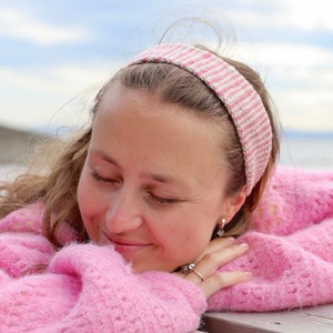 Puede incluir: Una mujer con el pelo largo y castaño que lleva una diadema a rayas rosa y blanca y un jersey rosa peludo. Está sonriendo y mirando hacia abajo.