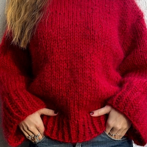 Könnte beinhalten: Ein leuchtend roter, grob gestrickter Pullover mit Rollkragen und ausgestellten Ärmeln. Der Pullover wird mit einer blauen Jeans kombiniert. Die Textur des Strickwerks ist deutlich sichtbar und erzeugt eine gemütliche und warme Ästhetik.