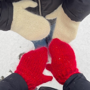 Könnte beinhalten: Draufsicht auf zwei Paar Fäustlinge. Zwei cremefarbene Strickfäustlinge werden zusammengehalten, und zwei leuchtend rote Strickfäustlinge werden zusammengehalten. Die Fäustlinge liegen auf einer schneebedeckten Oberfläche.