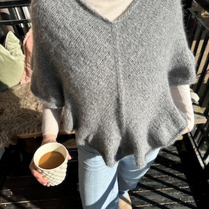 Puede incluir: Un poncho gris de manga corta con cuello en V. El poncho está hecho de un material suave y tejido. La persona lleva un cuello alto color crema y vaqueros azul claro. Se sostiene una taza blanca con un patrón texturizado en la mano.