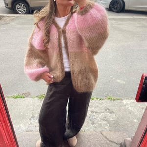Pode incluir: Um cardigan de malha rosa e castanho com decote em V e mangas compridas bufantes. O cardigan é aberto na frente e parece ser feito de um fio macio e texturizado. As mangas são de uma cor rosa mais clara, enquanto o corpo é uma mistura de rosa e castanho.