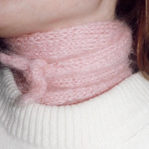 May include: A pink fuzzy knit scarf with a loose, knotted design. The scarf is worn around the neck of a person wearing a white sweater.