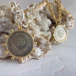 May include: Two gold necklaces with silver coin pendants. The pendants feature a detailed design with a crown and floral motifs. The necklaces are displayed on a white rock background.