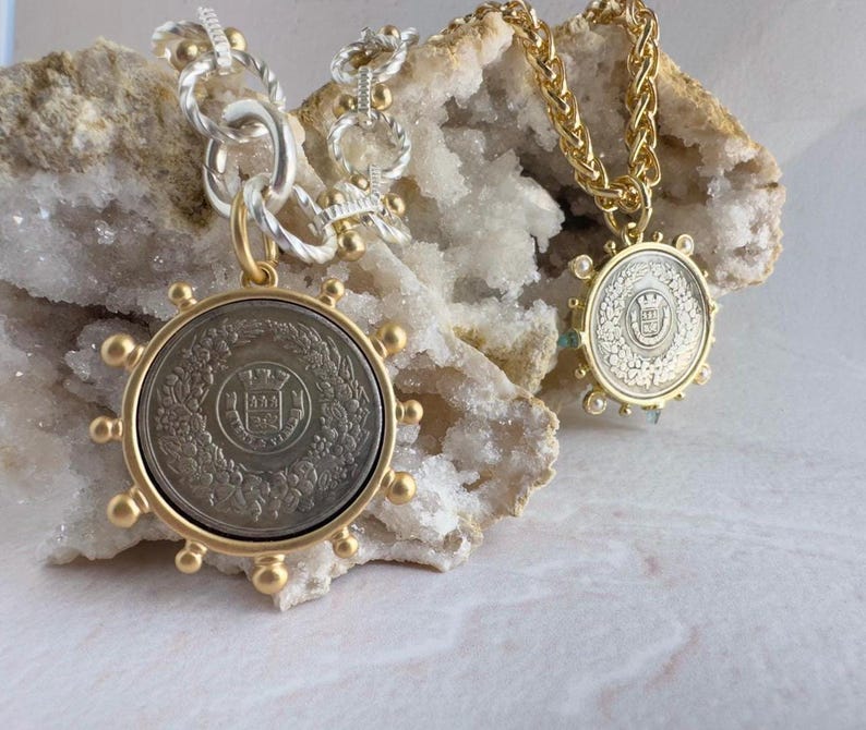 May include: Two gold necklaces with silver coin pendants. The pendants feature a detailed design with a crown and floral motifs. The necklaces are displayed on a white rock background.