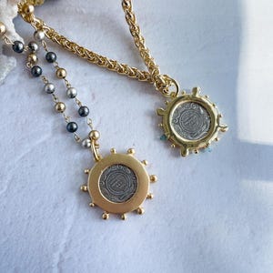 May include: Two gold necklaces with silver coin pendants. The pendants are surrounded by a decorative gold border. One necklace has a black and silver beaded chain, the other has a gold chain.