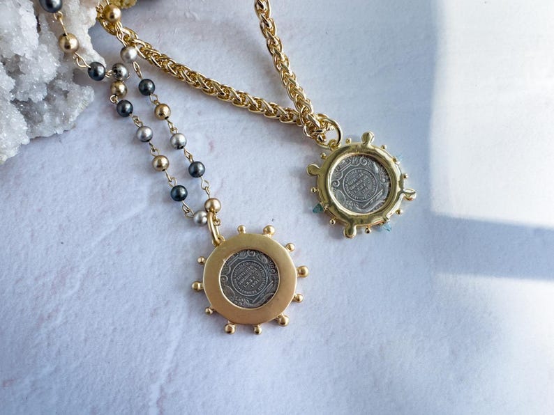 May include: Two gold necklaces with silver coin pendants. The pendants are surrounded by a decorative gold border. One necklace has a black and silver beaded chain, the other has a gold chain.