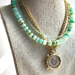 May include: A layered necklace with a gold chain and three strands of light green beads. The necklace has a large, round pendant with a gold design.