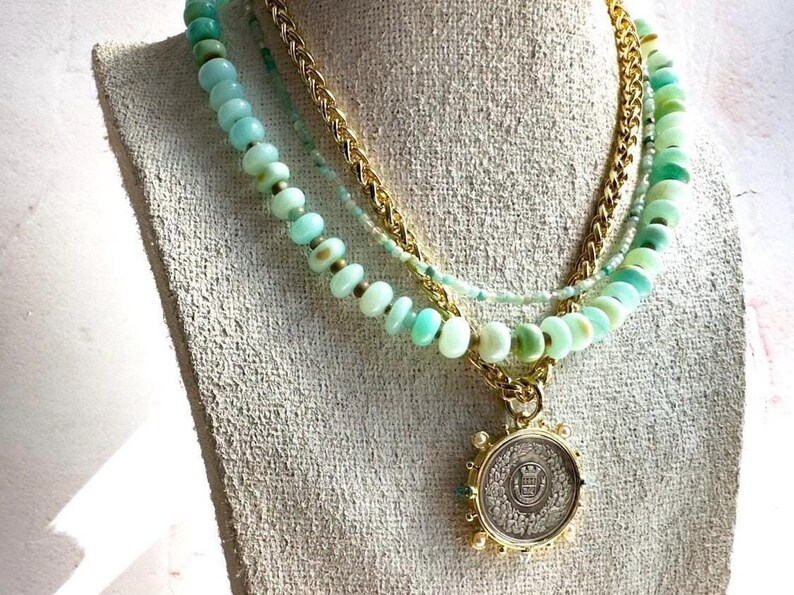 May include: A layered necklace with a gold chain and three strands of light green beads. The necklace has a large, round pendant with a gold design.