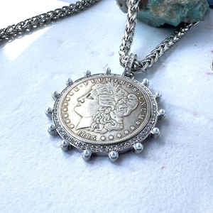 Könnte beinhalten: Eine silberfarbene Anhängerkette mit einer 1885 Morgan-Silberdollar-Münze in der Mitte. Die Münze ist von einem kreisförmigen Rand mit kleinen klaren Steinen umgeben und mit winzigen silbernen Kugeln am äußeren Rand verziert, hängend an einer dicken Kette.