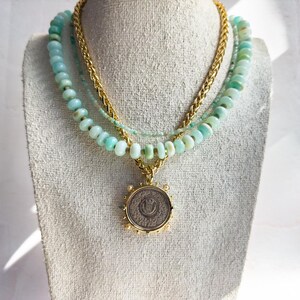 May include: A layered necklace with a gold chain and three strands of light blue opal beads. A gold pendant with a silver coin is hanging from the necklace.
