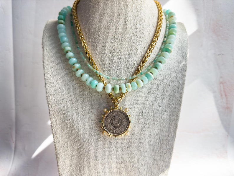 May include: A layered necklace with a gold chain and three strands of light blue opal beads. A gold pendant with a silver coin is hanging from the necklace.