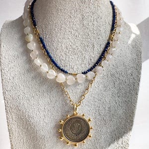May include: A layered necklace with a gold chain and a large, round pendant. The pendant has a detailed design and is surrounded by small gold beads. The necklace is also adorned with white beads and a blue beaded strand.