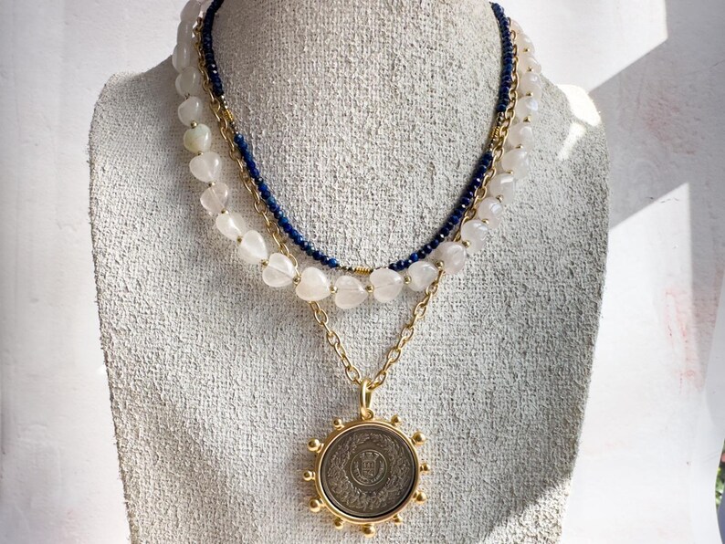 May include: A layered necklace with a gold chain and a large, round pendant. The pendant has a detailed design and is surrounded by small gold beads. The necklace is also adorned with white beads and a blue beaded strand.