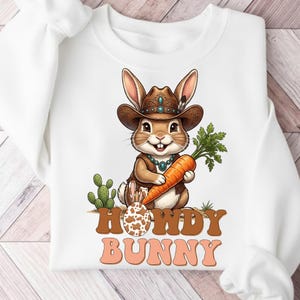May include: White sweatshirt featuring a cartoon bunny wearing a cowboy hat and vest, holding a carrot. The design includes the text "Howdy Bunny" in brown and orange. A cactus and a decorated egg are also part of the graphic.