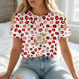 Op de afbeelding: Wit T-shirt met een patroon van rode, roze en gouden luipaardvlekken en harten. Een grote witte roos met gouden accenten is gecentreerd op de voorkant. Het shirt heeft korte mouwen en een ronde hals.