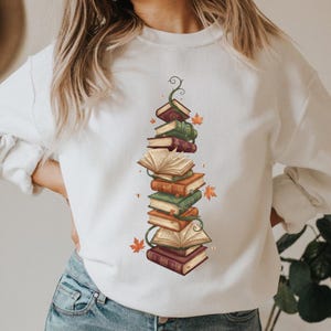 Puede incluir: Sudadera blanca con un diseño de una pila de libros con una vid y hojas de otoño. Los libros son de varios colores, incluyendo burdeos, verde y naranja. El diseño está centrado en la parte delantera de la sudadera.