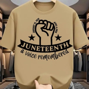 May include: A tan t-shirt with a black graphic of a raised fist, stars, and the text "Juneteenth a voice remembered." Dates January 15, 1929 and April 4, 1968 are included. The shirt has short sleeves and a crew neck.