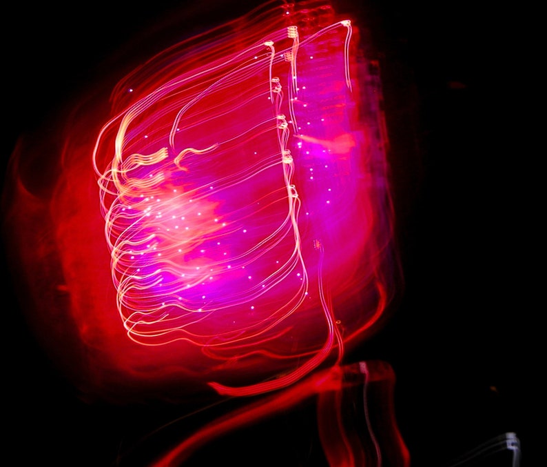 Op de afbeelding: Abstracte afbeelding met een zwarte achtergrond en een levendige weergave van rode en roze lichtstrepen. Het licht vormt een centrale, gloeiende vorm met delicate, witte punten, waardoor een dynamische en artistieke compositie ontstaat.