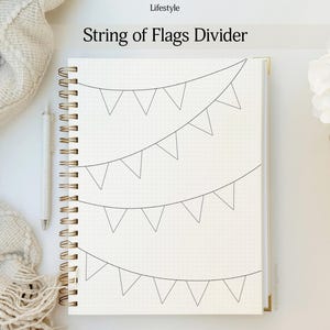 May include: A notebook with a gold spiral binding, featuring a dotted grid pattern and a hand-drawn string of flags design. The text "String of Flags Divider" is at the top. A pen rests on the left side, with a soft blanket and flowers in the background.
