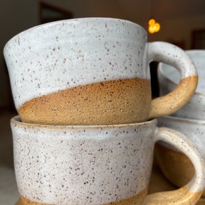 Puede incluir: Dos tazas de cerámica blanca con fondos y asas moteados de marrón. Las tazas están apiladas una encima de la otra.