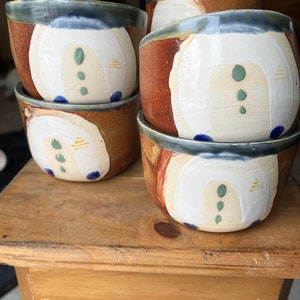May include: Four ceramic mugs with a white glaze and abstract brown, blue, and green designs. The mugs are stacked on top of each other on a wooden surface.