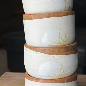 May include: A stack of four handmade ceramic bowls. The bowls are off-white with a brown rim and a white, leaf-like design with yellow accents. The bowls are stacked on top of each other.