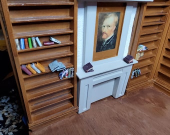 May include: A miniature wooden library set featuring tall shelves filled with tiny colorful books and a central light gray fireplace mantel. A framed portrait of a man with a reddish beard is mounted above the mantel, with two small books resting on the ledge.
