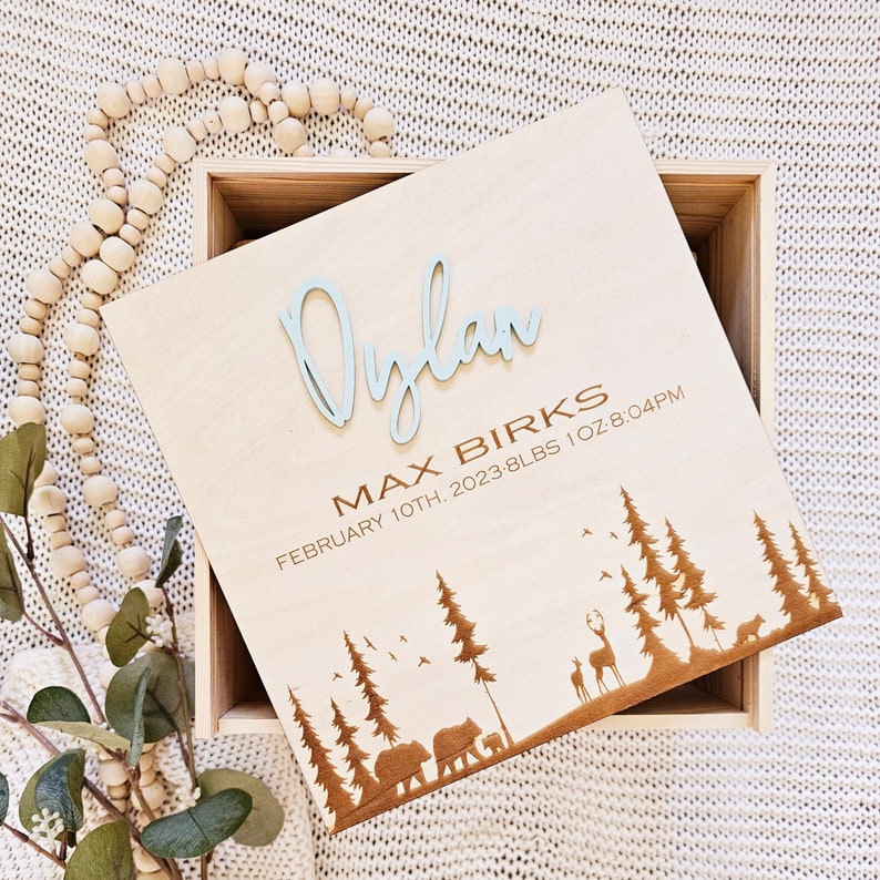 May include: Wooden keepsake box with a light blue engraved name "Dylan Max Birks" and the date "February 10th, 2023 - 8 lbs 1 oz 8:04 PM". The box has a woodgrain pattern and a laser-cut design of trees, bears, and deer.