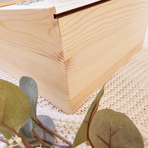 May include: A wooden box with a natural wood finish. The box is closed and the lid is slightly ajar. The box is sitting on a white knitted surface.
