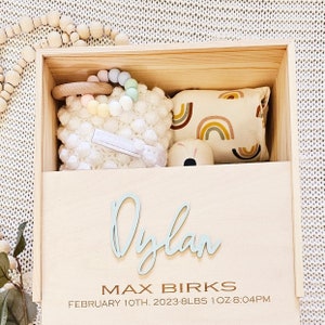 May include: Wooden keepsake box with a light blue inscription of the name "Dylan", the text "Max Birks", and the date "February 10th, 2023 - 8 lbs 1 oz 8:04 PM". The box contains a white crocheted stuffed animal, a rainbow-coloured teether, a wooden toy, and a small pillow with a rainbow pattern.