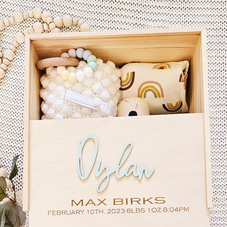 May include: Wooden keepsake box with a light blue inscription of the name "Dylan", the text "Max Birks", and the date "February 10th, 2023 - 8 lbs 1 oz 8:04 PM". The box contains a white crocheted stuffed animal, a rainbow-coloured teether, a wooden toy, and a small pillow with a rainbow pattern.