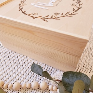 May include: A natural wood box with a floral design and the word "Love" engraved on the top. The box is sitting on a white knitted surface with a string of wooden beads and a eucalyptus branch.