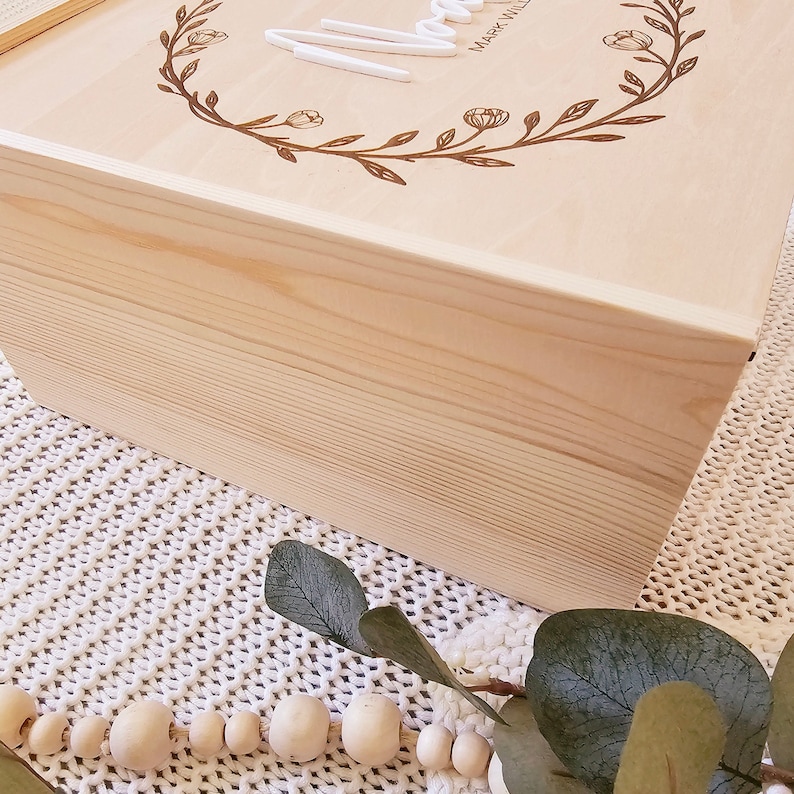 May include: A natural wood box with a floral design and the word "Love" engraved on the top. The box is sitting on a white knitted surface with a string of wooden beads and a eucalyptus branch.