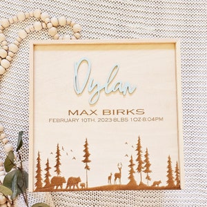 May include: A wooden sign with a light blue name "Dylan" and the text "Max Birks, February 10th, 2023- 8lbs 1oz 8:04PM". The sign has a brown silhouette of a forest scene with trees, deer, and bears.