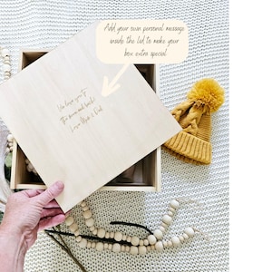 May include: A wooden box with a lid that can be personalised with a message. The lid is open and the inside of the box is visible. The box is sitting on a white knitted surface. There is a yellow knitted hat with a pom-pom on top of the box.