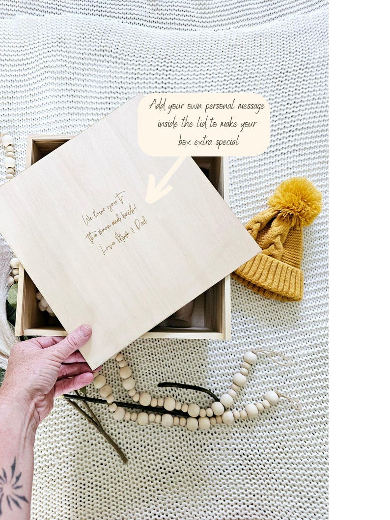May include: A wooden box with a lid that can be personalised with a message. The lid is open and the inside of the box is visible. The box is sitting on a white knitted surface. There is a yellow knitted hat with a pom-pom on top of the box.