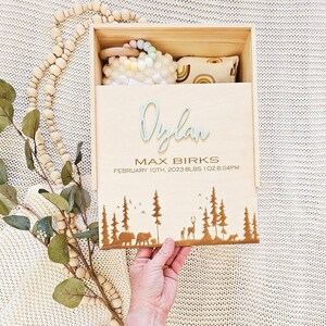 May include: Wooden keepsake box with a light blue engraved name "Dylan" and the text "Max Birks, February 10th, 2023, 8 lbs 1 oz, 8:04 PM". The box has a forest scene with trees, birds, and deer engraved on the bottom.