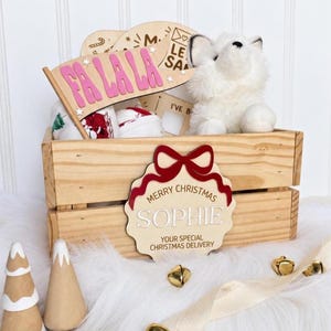 Könnte beinhalten: Eine Holzkiste mit einem personalisierten Schild, einer roten Schleife und dem Text Merry Christmas Sophie Your Special Christmas Delivery. Darin befinden sich ein weißer Plüschfuchs, ein Holzwimpel mit rosa FA LA LA Schriftzug und Stoffartikel.