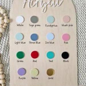 May include: A wooden sign with the word "Acrylic" at the top. The sign has 12 colour swatches, each with a name below it. The colours are: white, sage green, eucalyptus, blush pink, light blue, stone blue, ice blue, pink, green, red, teal, black, purple, yellow, and gold.
