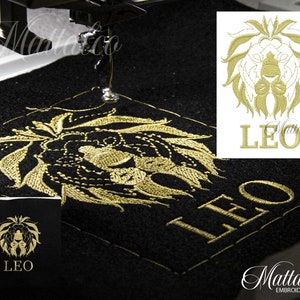 Può includere: Designo ricamato in oro di una testa di leone con la parola "LEO" sotto. Il design è cucito su un tessuto nero con una macchina da cucire.