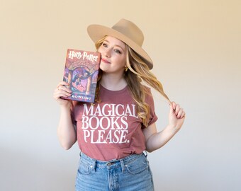 May include: A woman wearing a brown hat and a maroon t-shirt with the text "MAGICAL BOOKS PLEASE." is holding a copy of the book "Harry Potter and the Sorcerer's Stone" by J.K. Rowling.