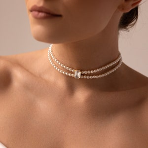May include: Double-strand pearl choker necklace with a rectangular, clear crystal pendant set in gold. The necklace is worn around the neck, showcasing the delicate pearls and the sparkling crystal. The jewelry is elegant and suitable for special occasions.