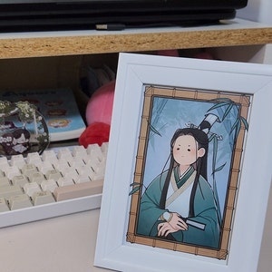 Op de afbeelding: Een wit ingelijste print van een cartoonfiguur met lang zwart haar, gekleed in een groene en witte robe, zittend in een bamboebos. Het figuur heeft een lichtblauwe achtergrond en is omringd door groene bamboe.