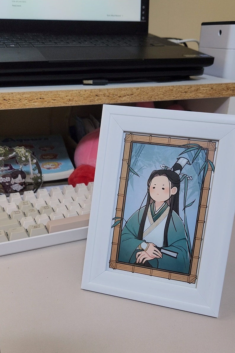 Op de afbeelding: Een wit ingelijste print van een cartoonfiguur met lang zwart haar, gekleed in een groene en witte robe, zittend in een bamboebos. Het figuur heeft een lichtblauwe achtergrond en is omringd door groene bamboe.