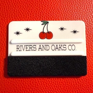 May include: A white squeegee with a black scrubbing pad at the bottom. The top of the squeegee features a cherry design and the text "RIVERS AND OAKS CO." is printed below the cherry design. The background is red.